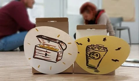 Zwei gestaltete Feedbackboxen auf einem Tisch, im Hintergrund arbeiten Menschen.
